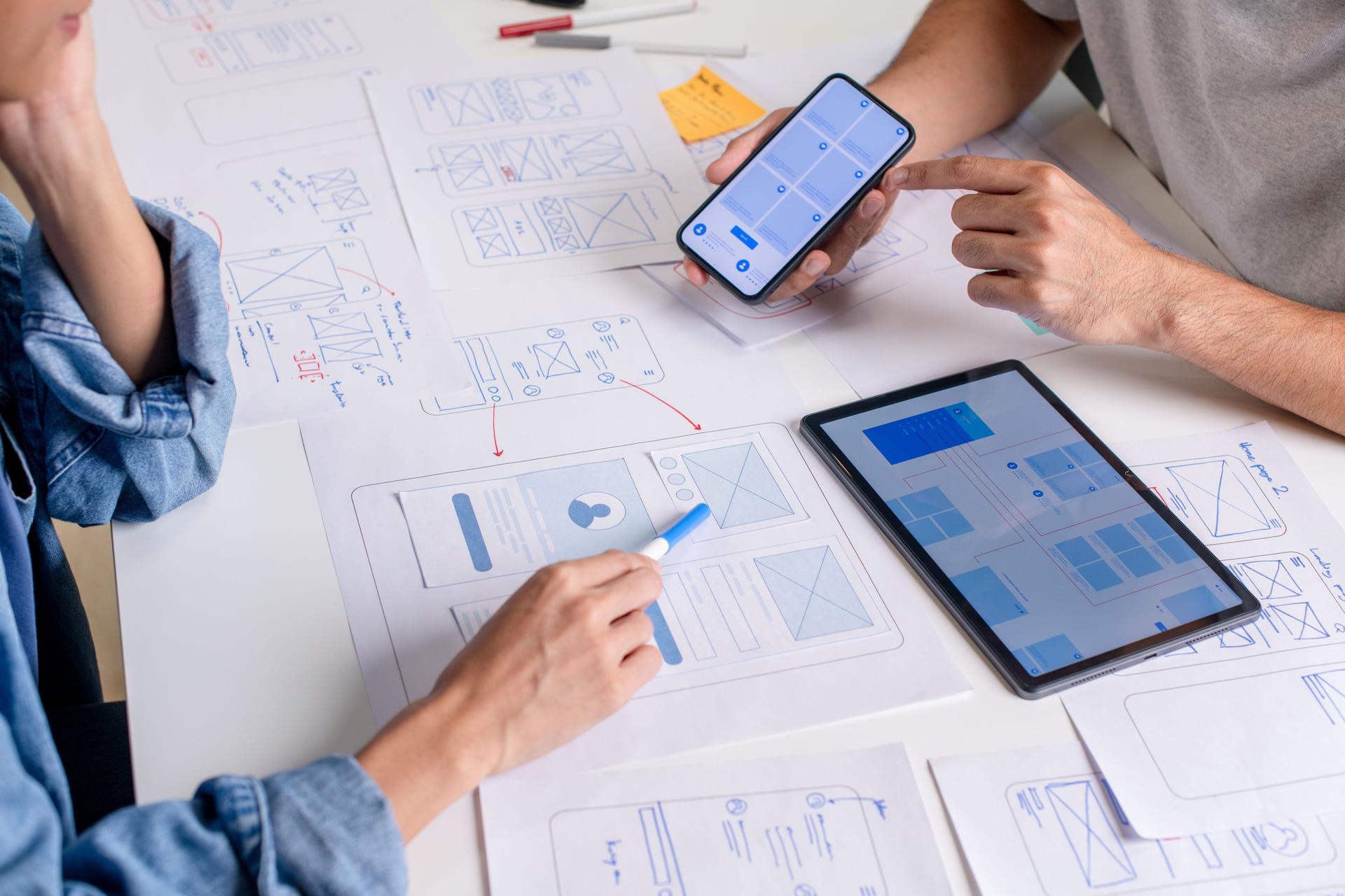 UX/UI designer testing prototype on a phone, discussing and brainstorming on wireframes for a website and mobile app prototype, surrounded by sketches of user flow and design tools, in the concept of website and mobile application design concept.